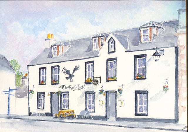 The Eagle Hotel in Lauder, a white two-storey building with dormer windows, a painted eagle on the wall, hanging baskets and a picnic bench.