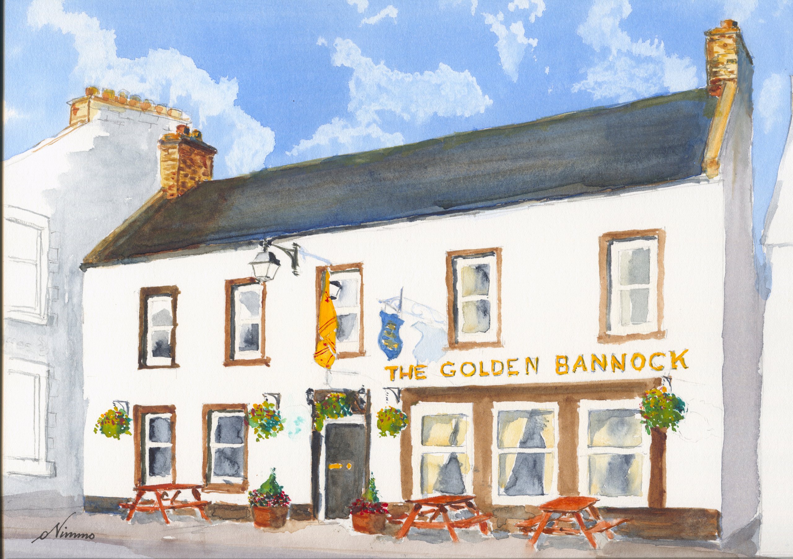 The Golden Bannock in Lauder, a white pub with a dark roof, its name in gold letters, a yellow flag, hanging baskets and red picnic tables under a blue sky.