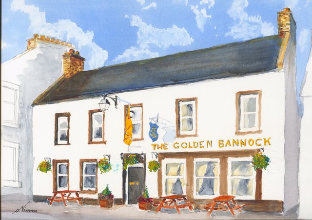 The Golden Bannock in Lauder, a white pub with a dark roof, its name in gold letters, a yellow flag, hanging baskets and red picnic tables under a blue sky.